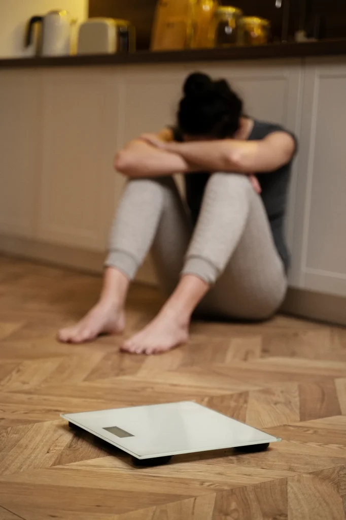 Persona sentada en el suelo con la cabeza entre las rodillas junto a una báscula