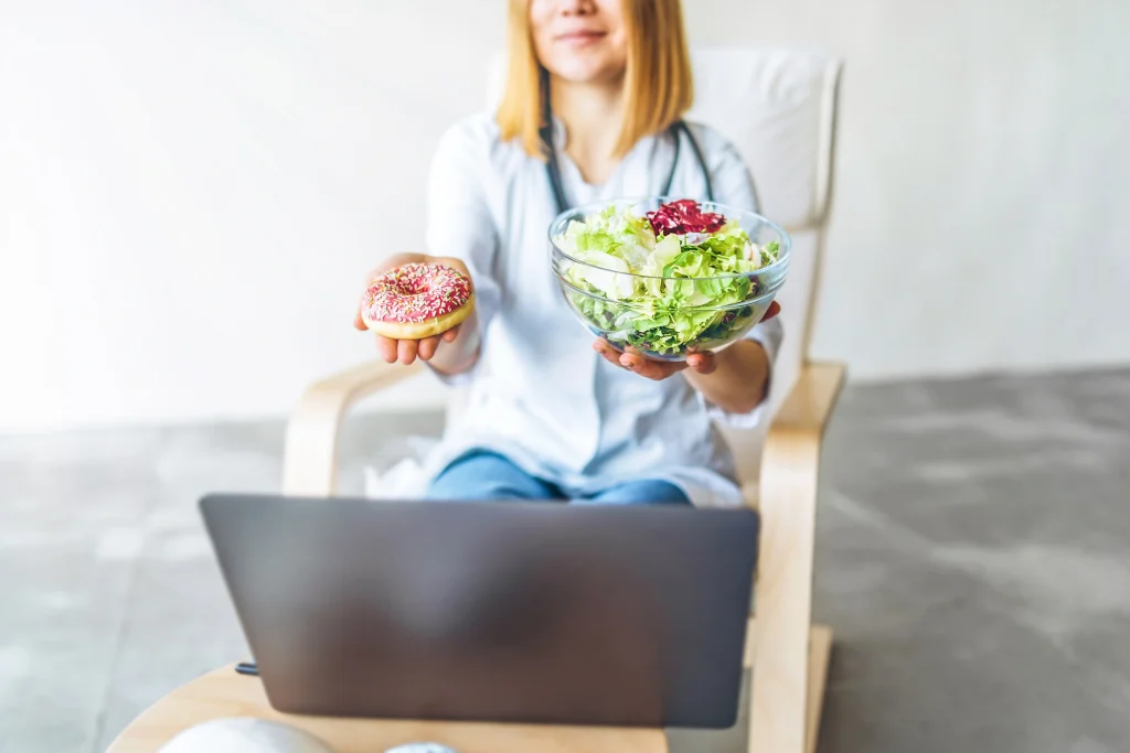 Nutricionista con bata sosteniendo una ensalada y un dónut frente a un portátil